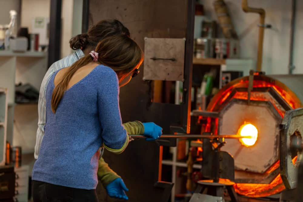 AdamAaronson-GlassBlowing Two people are shaping glass with a rod in front of a hot furnace in a workshop.