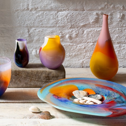 AdamAaronsonVases Colorful glass vases and a decorative plate with shells on a light wooden surface.