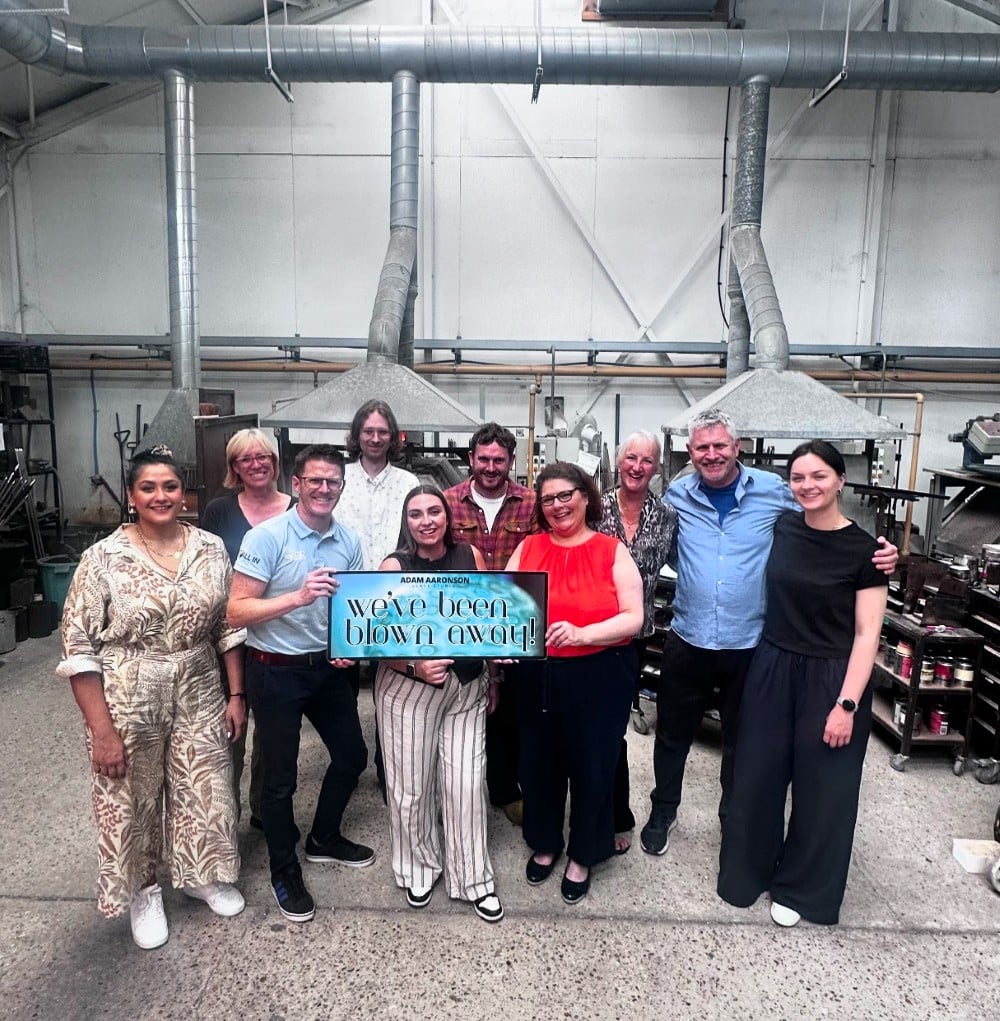 CorporateGroupBooking A group of people smiling and holding a sign that says "we've been blown away!" in a workshop.