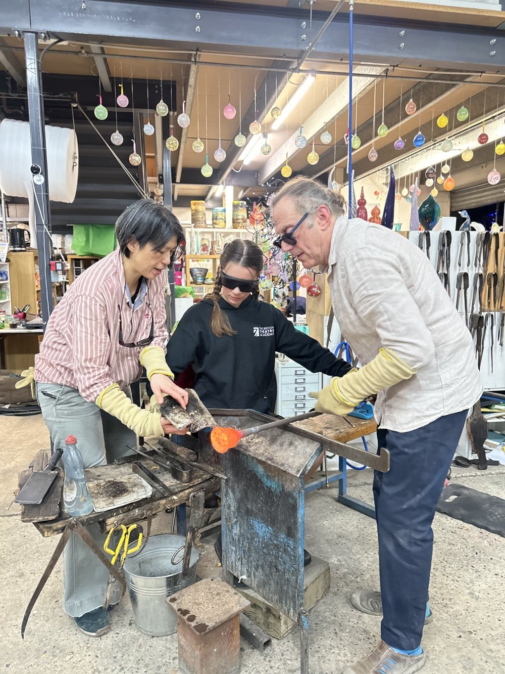 ExploreOurCourses Three people working together on a glassblowing project in a colorful workshop.